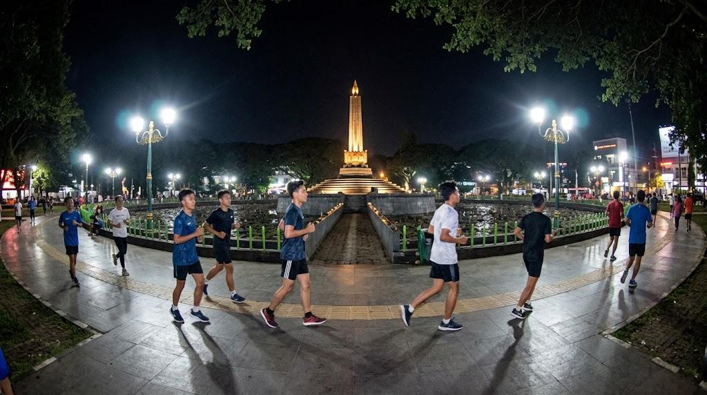 Anak muda jogging pada malam hari di area olahraga dengan pakaian olahraga dan lampu penerangan.
